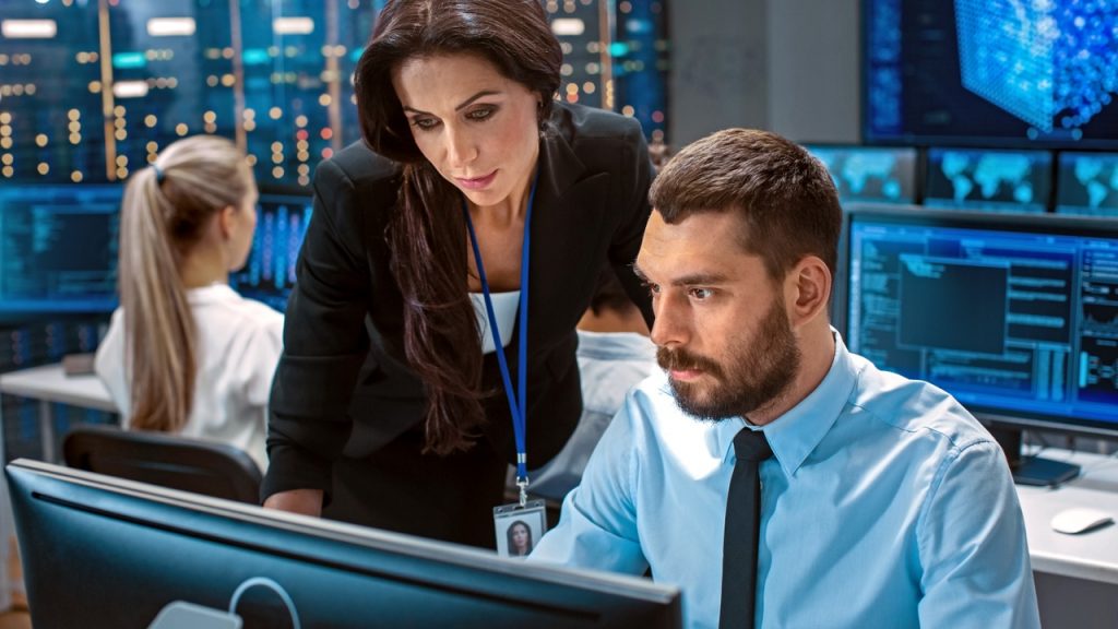 Female Chief Computer Engineer Consults Male Neural Network Architect. They Work in a Crowded Office on a Neural Network/ Artificial Intelligence Project. Office Space Has Data Server