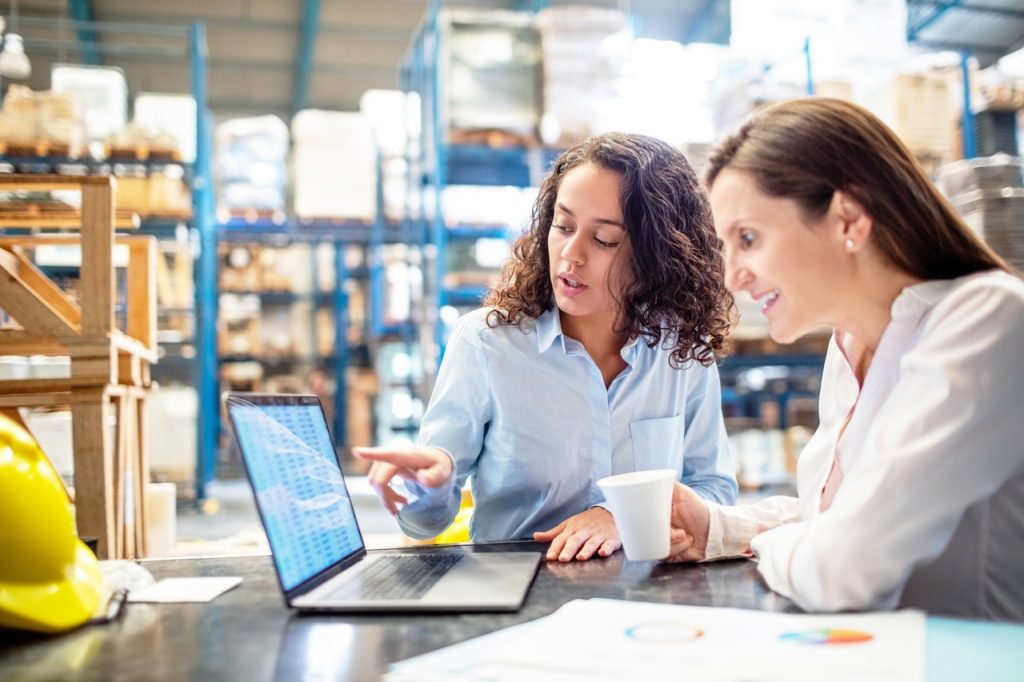 Young woman showing something on laptop to manager in factory warehouse. Two business women working together on laptop, checking the inventory.