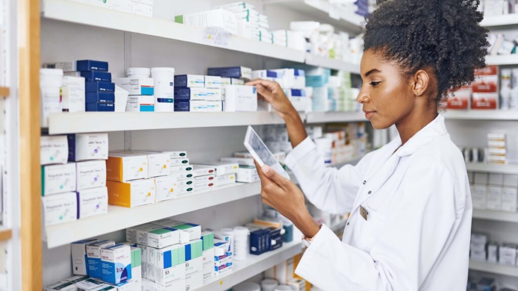 Photo of a young black woman pharmacist checking stock on pharmacy shelves. She has a tablet in her hand.