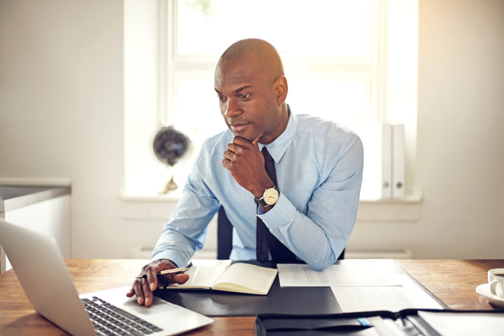 Young African business executive wearing a shirt and tie sitting at his desk in an office working online with a laptop