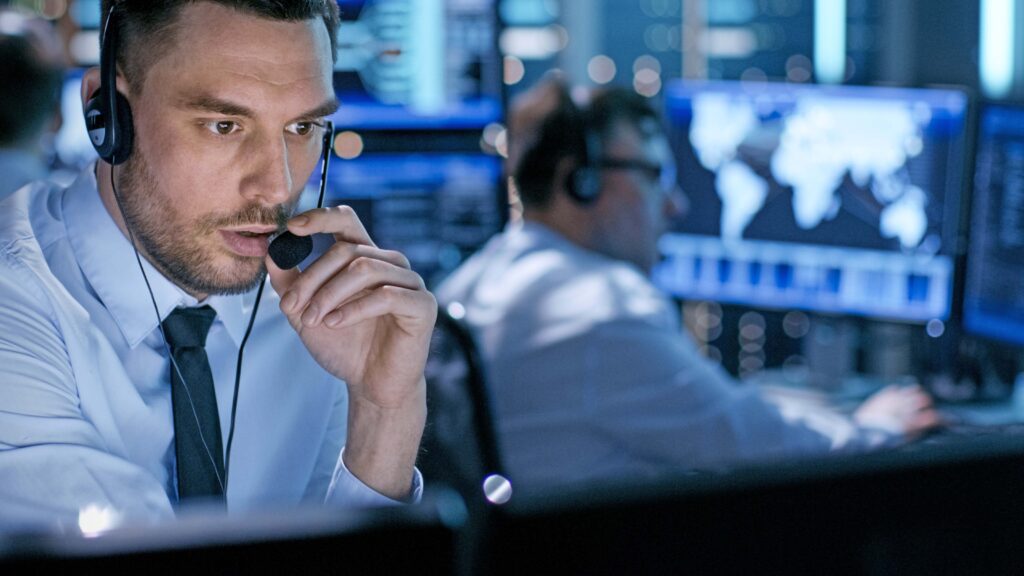 A man in format clothes works in front of a computer and talks into a headset microphone.