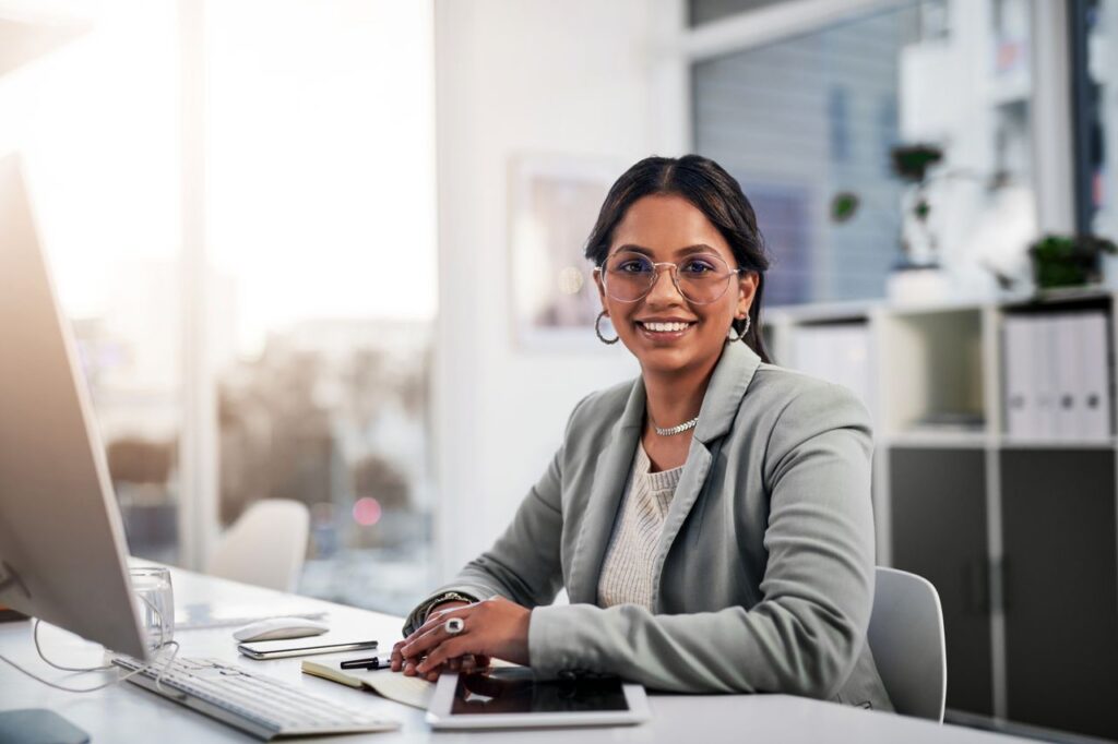 Woman with dark hair seated at a desk in a bright office, smiling at the camera. They wear glasses, a light blazer over a white top, and jewelry. The desk holds a computer monitor, keyboard, notepad, pen, smartphone, and tablet. Shelves with binders and plants are visible in the background, with natural light streaming through large windows.