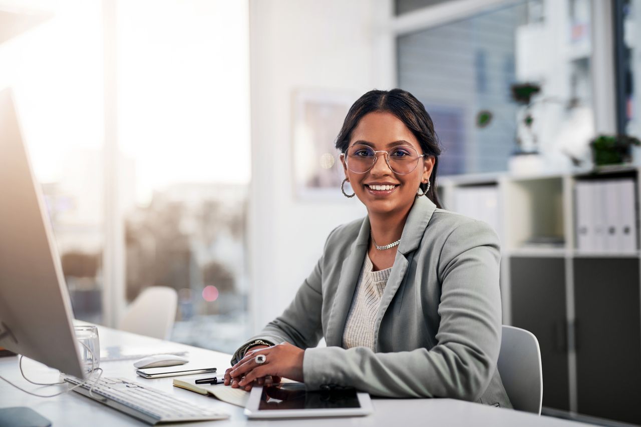 Woman with dark hair seated at a desk in a bright office, smiling at the camera. They wear glasses, a light blazer over a white top, and jewelry. The desk holds a computer monitor, keyboard, notepad, pen, smartphone, and tablet. Shelves with binders and plants are visible in the background, with natural light streaming through large windows.