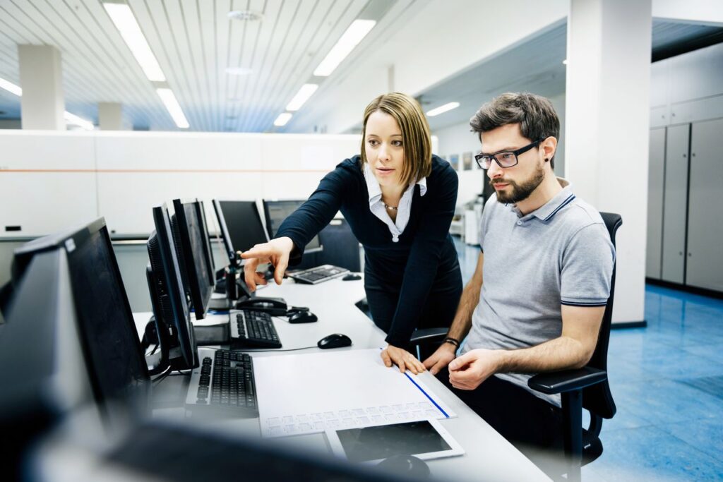 Two IT professionals in a big control room of a factory talking.