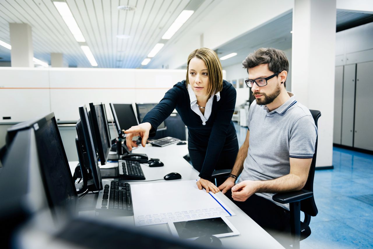 Two IT professionals in a big control room of a factory talking.