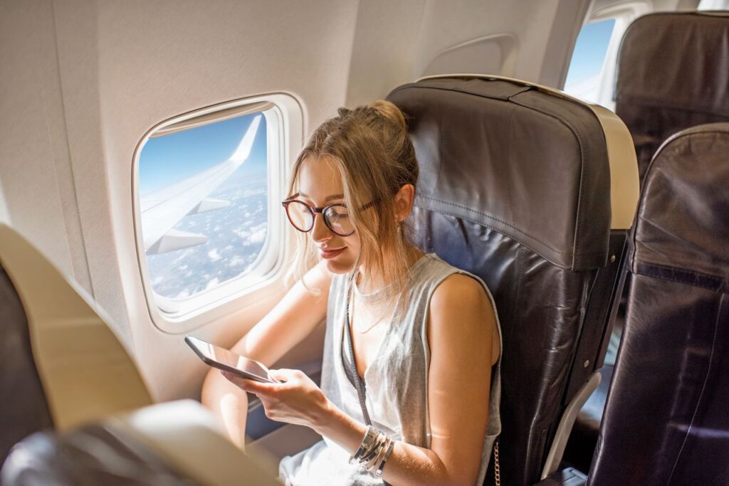 woman looking at her phone while seated in an airplane next to a window on a sunny day