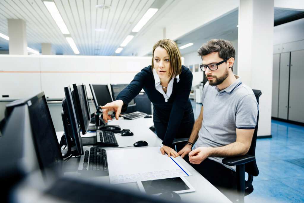 Two IT professionals in a big control room of a factory talking.