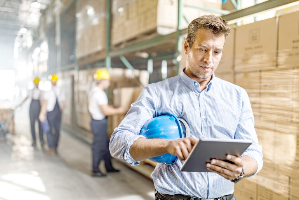 Mid adult foreman surfing the Internet on touchpad in a distribution warehouse. There are people in the background.