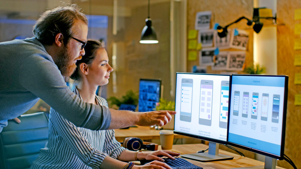 Female UX Architect Has Discussion with Male Design Engineer, They Work on Mobile Application Late at Night, She Drinks Coffee. In the Background Wall with Project Sticky Notes.