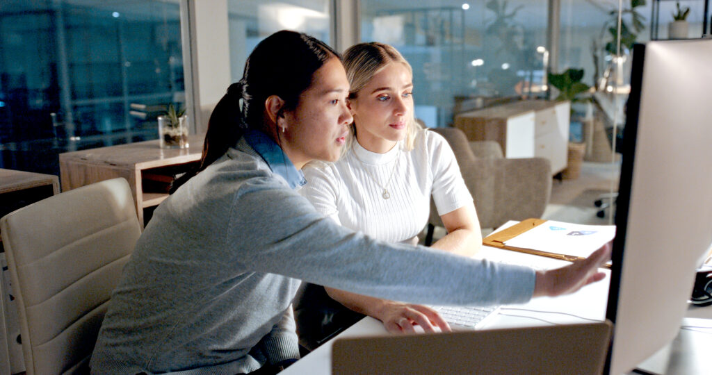 A person seated at a desk points at a computer monitor while another person sits beside them, both collaborating on work in a modern office setting with laptops, papers, and office furniture visible in the background.