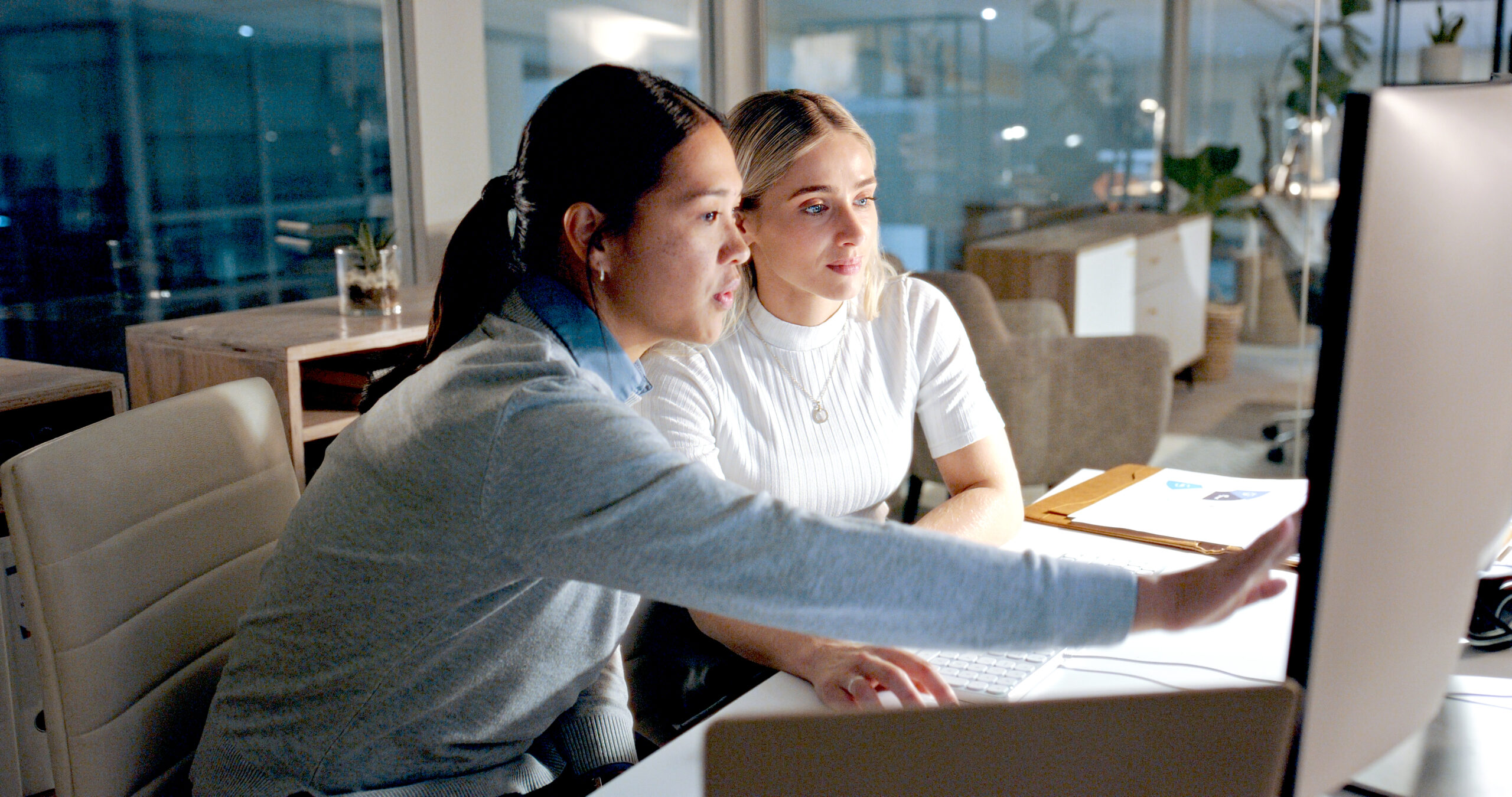A person seated at a desk points at a computer monitor while another person sits beside them, both collaborating on work in a modern office setting with laptops, papers, and office furniture visible in the background.