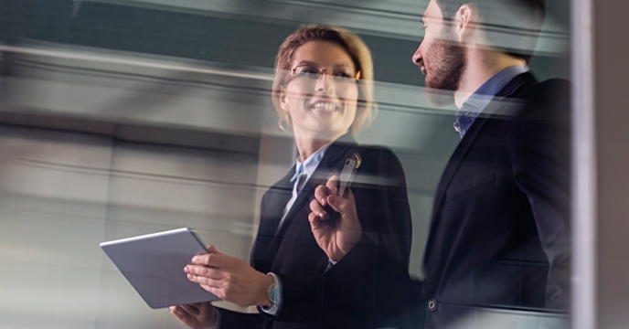 Two business professionals walking through a modern office corridor, holding a tablet and a smartphone, seen through glass with motion blur, suggesting fast‑moving decision‑making and coordination in a digital enterprise environment.