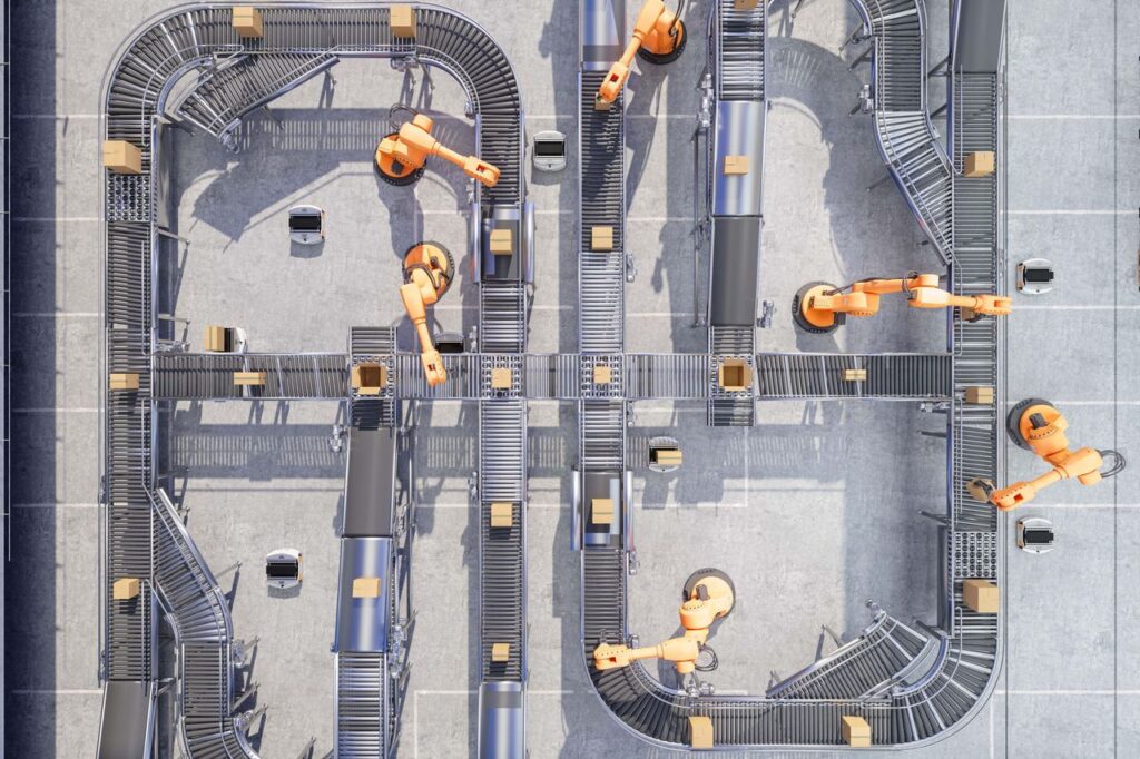 Overhead view of an automated warehouse conveyor system with multiple robotic arms sorting small boxes along intersecting belt lines.