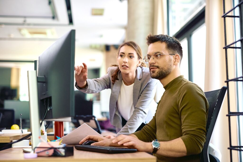 Deux personnes travaillent à un bureau dans un espace de bureau, devant un grand écran, tandis que l’une pointe l’écran et que l’autre utilise un clavier.